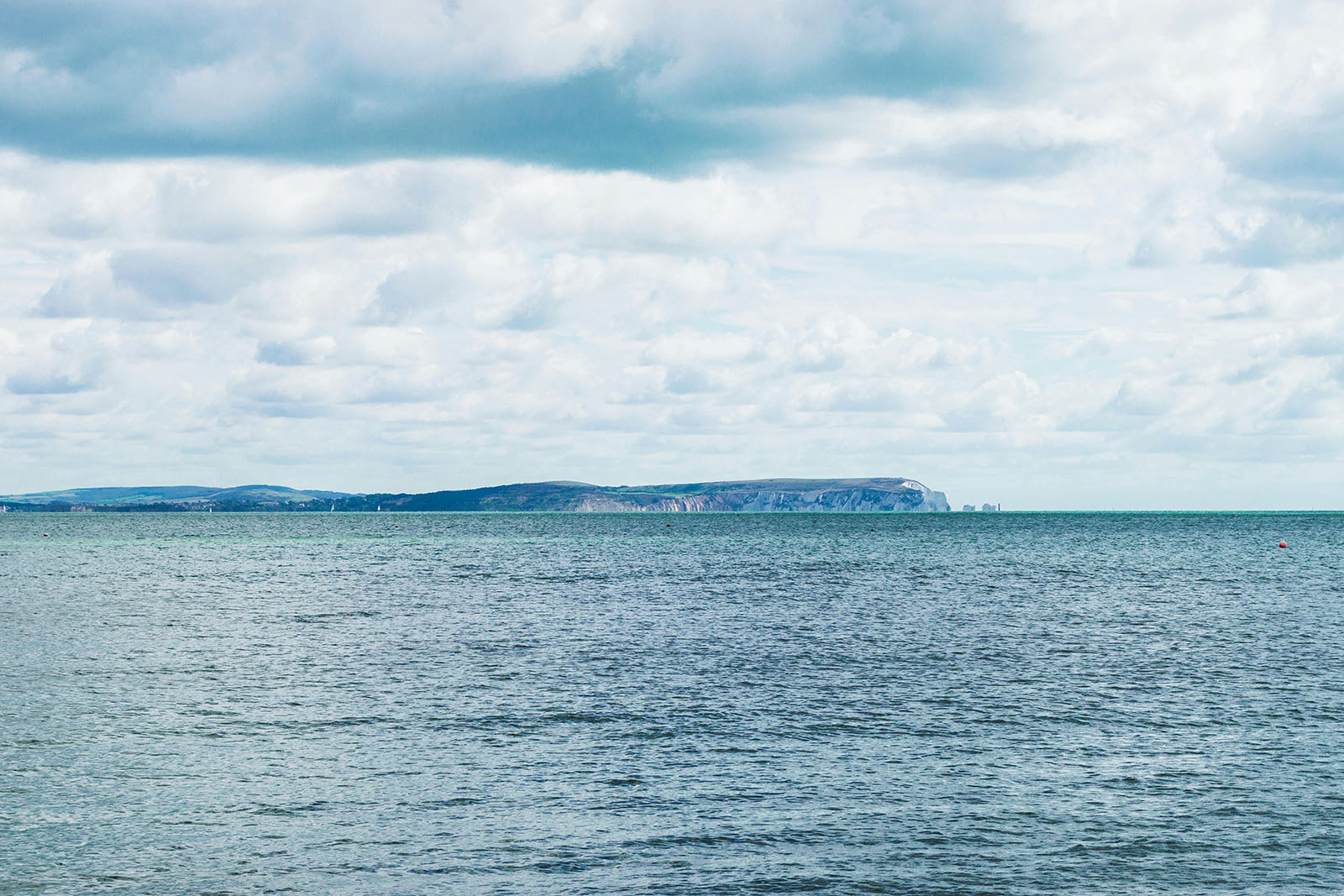 A wide seascape view of the Isle of Wight Needles in the distance beneath a cloudy sky, with calm blue-grey water in the foreground.